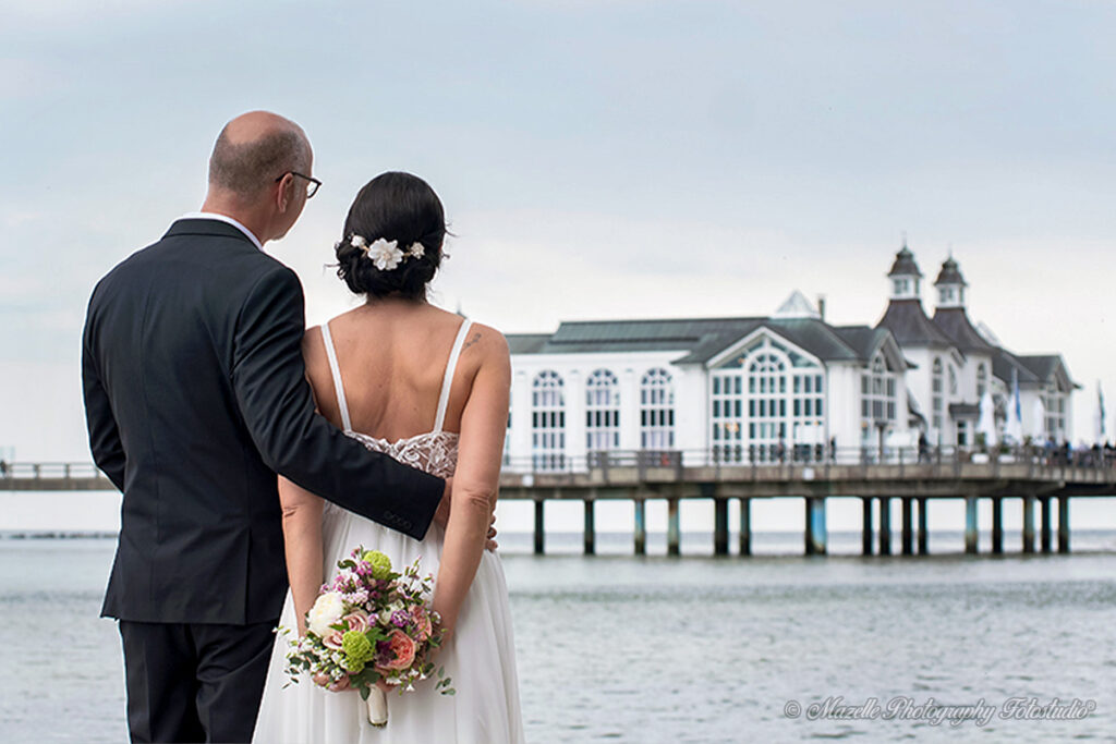 Nach der Trauung im historischen Trausaal der Seebrücke Sellin steht das Brautpaar am Strand und blickt gemeinsam auf die Kulisse ihrer Liebe. Die Braut hält ihren Brautstrauß hinter dem Rücken, während sich Licht, Meer und Emotionen zu einem stillen Moment vereinen. Fotografisch begleitet vom Hochzeitsfotografen des Mazelle Photography Fotostudio® – mit künstlerischem Feingefühl und emotionaler Tiefe festgehalten.