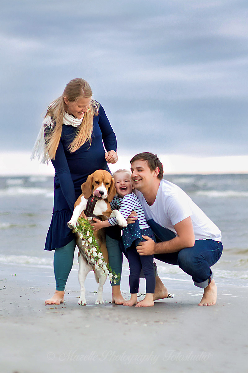 Eine Familie spaziert entspannt am Strand der Ostsee – begleitet von ihrem Kind und Hund. Die Frau trägt einen Strauß Wildblumen in der Hand, während die Küstenbrise und das Licht des Meeres den Moment umrahmen. Aufgenommen durch die Familienfotografie vom Mazelle Photography Fotostudio®, zeigt dieses Bild die stille Schönheit des Alltags, die Nähe zwischen Mensch und Tier und die Magie gemeinsamer Augenblicke in der Natur.