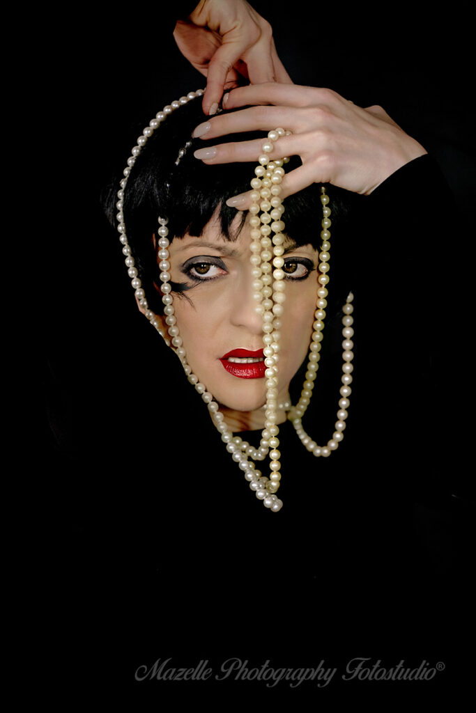 Ein einmaliges Portraitshooting von Nathalie im Mazelle Photography Fotostudio®. Vor schwarzem Hintergrund zeigt sich nur ihr Gesicht, über das eine elegante Perlenkette gelegt ist. Diese künstlerische Aufnahme entstand in Warschau, Polen, und spiegelt die internationale Ausrichtung unserer Portraitfotografie wider. Die Arbeiten von Mazelle Photography erscheinen regelmäßig in renommierten Zeitschriften und Magazinen und stehen für emotionale, authentische und hochwertige Fotokunst