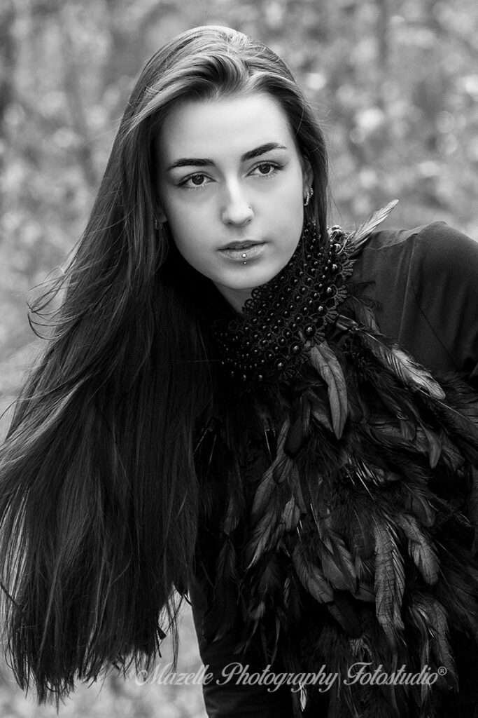 Künstlerisches Schwarz‑Weiß‑Portrait von Sophia im Krummen Wald bei Gryfino, Polen. Sie blickt hinter den charakteristisch gekrümmten Bäumen hervor – eine Aufnahme voller Ausstrahlung und stiller Eleganz. Das Portrait entstand im Rahmen eines besonderen Fotoshootings von Mazelle Photography Fotostudio®, das sich durch emotionale Tiefe und internationale Qualität auszeichnet.