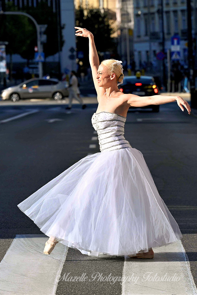 Künstlerisches Portraitshooting mit Ballerina Nathalie auf einer Straßenkreuzung in Warschau, Polen. Inmitten des normalen Verkehrs tanzt sie elegant, während Autos vorbeifahren und Fußgänger die Straße überqueren. Passanten beobachten die Szene und filmen mit ihren Handys – ein urbanes, ausdrucksstarkes Fotoshooting voller Energie und Authentizität, festgehalten von Mazelle Photography Fotostudio®.