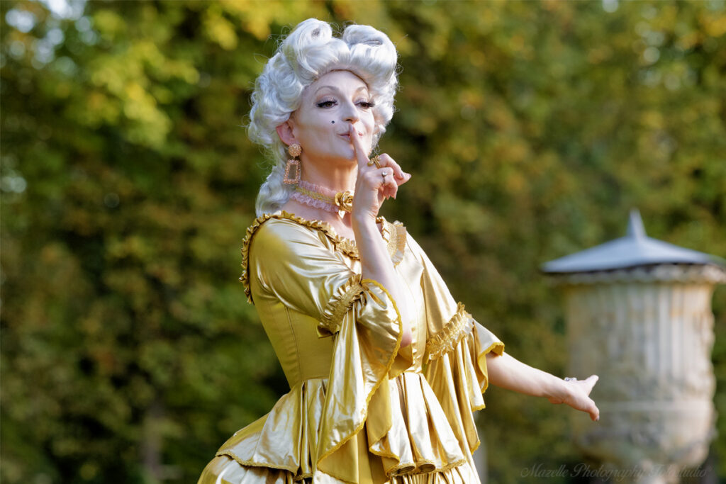 Nathalie hat sich im Schlosspark Wilanów in Warschau, Polen, als Marie Antoinette verkleidet. Sie trägt ein goldenes Kleid, weiße Haare und ein blass geschminktes Gesicht – ganz im Stil der Damen der Barockzeit. Die historische Kulisse des Schlosses Wilanów verstärkt die Authentizität und Eleganz der Szene und verleiht der Aufnahme eine besondere Tiefe.