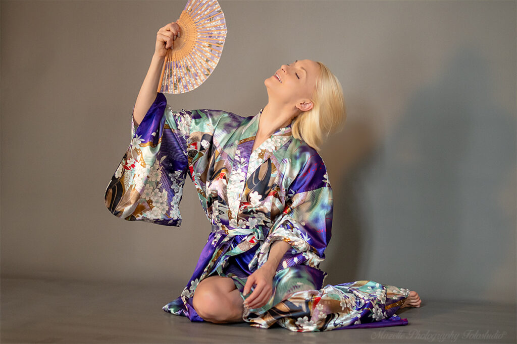 Nathalie trägt einen japanischen Kimono und spielt mit einem Fächer in der Luft, während sie auf dem Boden sitzt. Diese Szene wurde im Studio in Warschau, Polen, von den Porträtfotograf:innen des Mazelle Photography Fotostudio® aufgenommen. Die Aufnahme verbindet kulturelle Eleganz mit künstlerischer Porträtfotografie und zeigt die kreative Handschrift des Studios.
