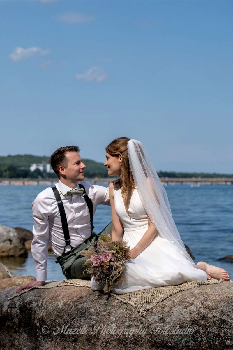 Ein frisch vermähltes Brautpaar sitzt auf einem Stein in der Ostsee vor Binz und schaut sich tief in die Augen – ein Moment voller Ruhe, Nähe und Verbundenheit. Das Hochzeitsfotoshooting fand direkt nach der Trauung im Müther-Turm statt und wurde exklusiv begleitet von den Hochzeitsfotografen des Mazelle Photography Fotostudio®. Die Ostsee, das Licht und die Natürlichkeit des Augenblicks verschmelzen zu einem Bild, das mehr erzählt als Worte.