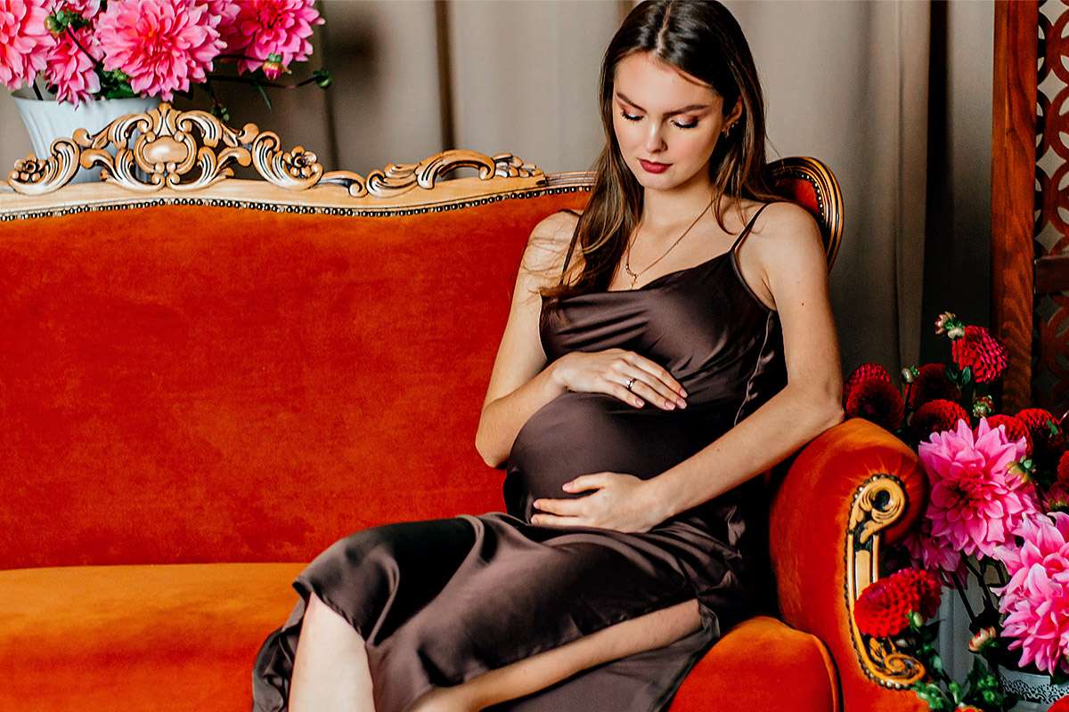 Eine schwangere Frau sitzt entspannt auf einem roten Sofa und streicht sanft über ihren Babybauch. Die ruhige, intime Atmosphäre zeigt die Vorfreude, Nähe und Verbundenheit mit dem ungeborenen Kind.