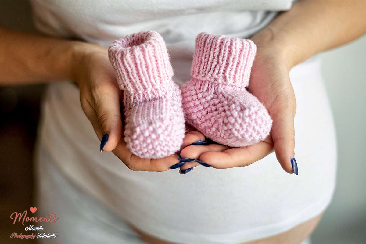 Ein liebevolles Schwangerschaftsportrait einer werdenden Mutter, die vor ihrem Babybauch ein Paar rosafarbene, handgestrickte Babyschuhe hält. Die Szene entsteht in der vertrauten Umgebung ihres eigenen Zuhauses – ruhig, entspannt und ohne Zeitdruck.