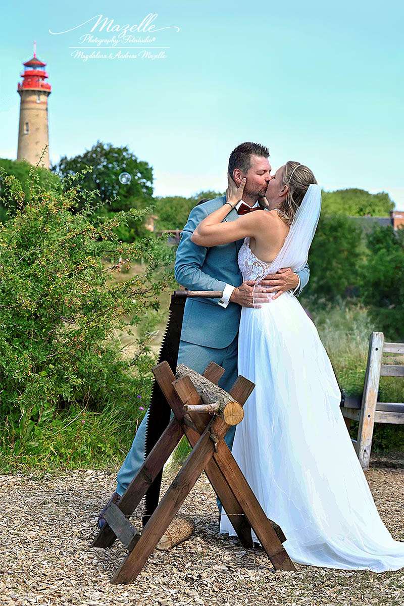 Das Brautpaar nach dem traditionellen Sägen des Baumstamms, festgehalten in einem echten Moment voller Nähe und Freude. Im Hintergrund erhebt sich der Leuchtturm von Kap Arkona, der die Szene trägt, ohne sie zu überlagern. Eine klare, zeitlose Aufnahme mit zwei Perspektiven und doppelter Tiefe – fotografiert von Mazelle Photography Fotostudio®.