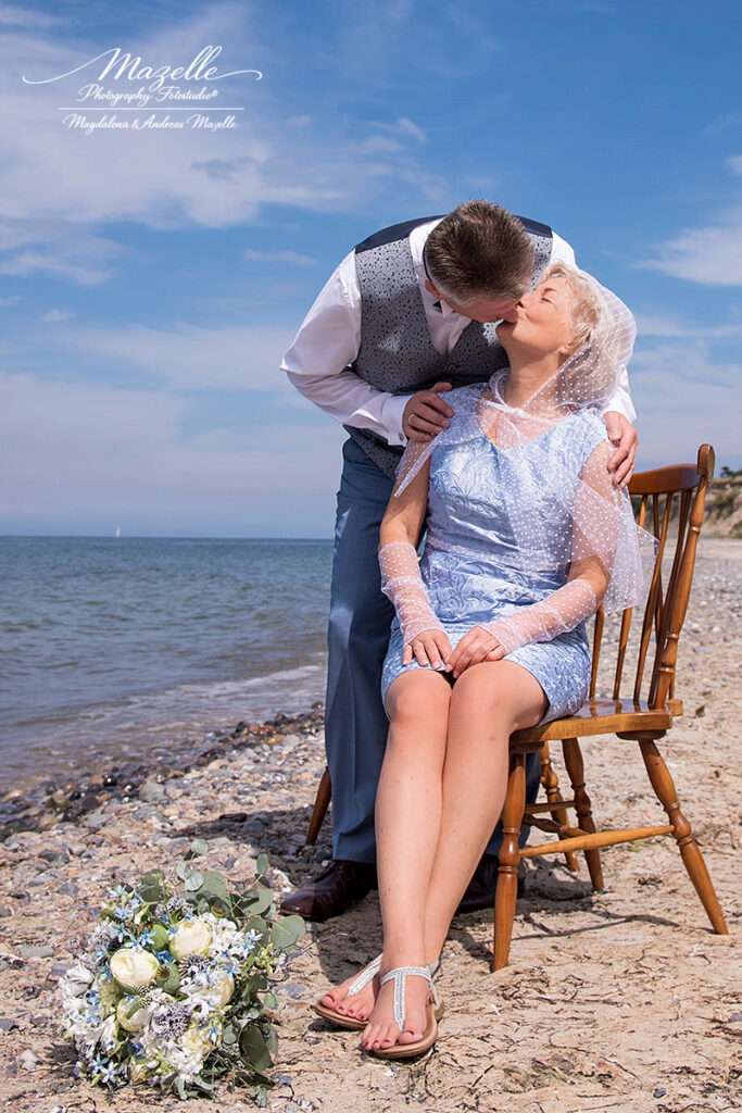 Am Strand der Krepnitzer Heide sitzt die Braut auf einem Stuhl, während der Bräutigam sich von hinten über sie beugt und ihr einen liebevollen Kuss schenkt – ein stiller, warmer Moment zwischen Ostsee, Wind und weitem Himmel.