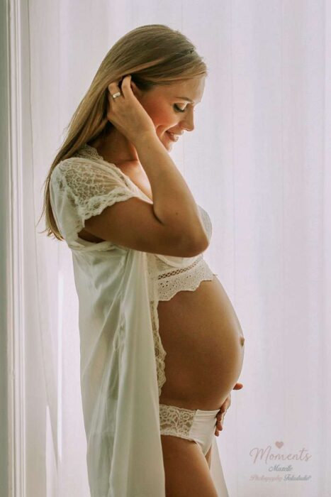 Eine schwangere Frau steht vor dem Fenster, streichelt liebevoll ihren Babybauch und blickt in einen Spiegel, der im Bild nicht sichtbar ist. Dieses Motiv der Babybauch- und Schwangerschaftsfotografie von Mazelle Photography Fotostudio® auf Moments.mazelle.de vereint Natürlichkeit, Eleganz und Emotion und zeigt die besondere Lebensphase werdender Mütter mit künstlerischer Tiefe